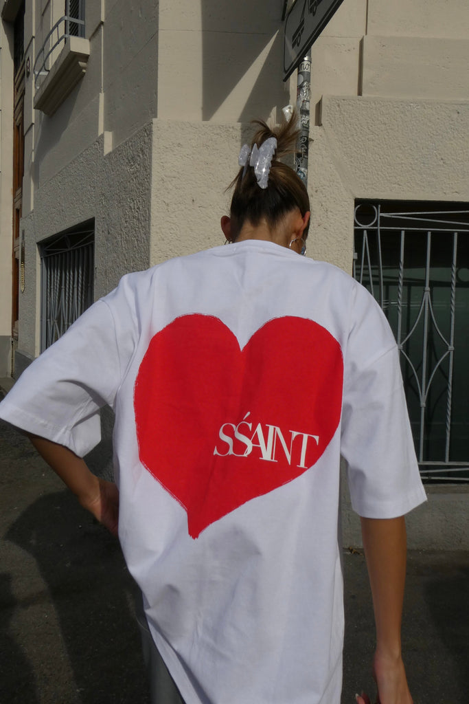 Person wearing a white t-shirt with a red heart and 'SS'ANT' logo on the back, standing on a street.